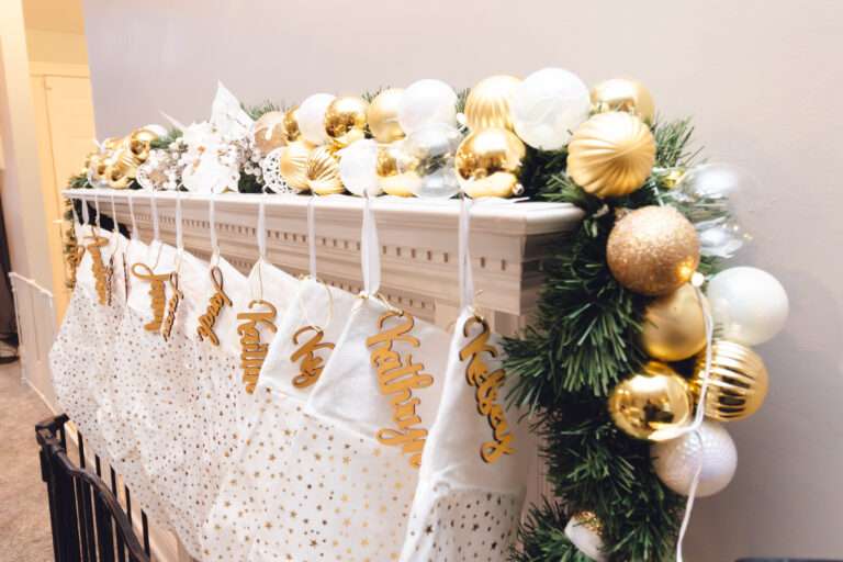 A mantle with some greenery, an abundance of gold, white, and clear ornaments, and many white stockings hung with gold decorations.