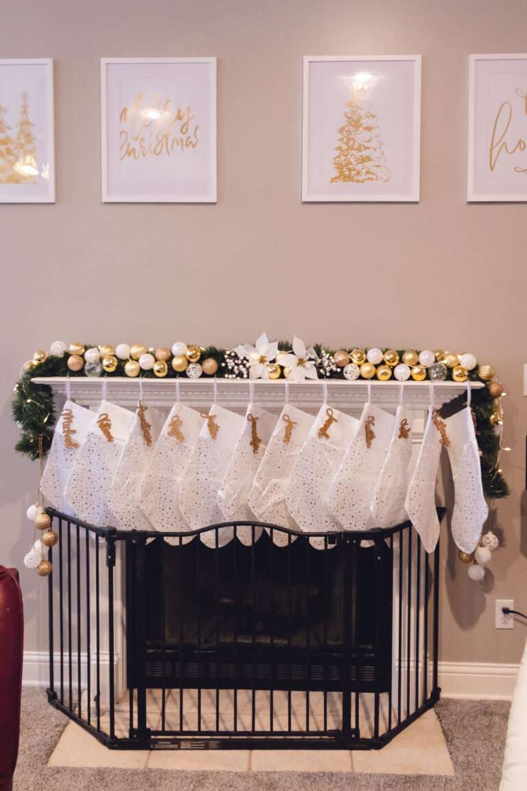 A mantle with some greenery, an abundance of gold, white, and clear ornaments, and many white stockings hung with gold decorations. Above the mantel are 4 white picture frames with gold holiday prints, and below the mantel, there is a black fireplace surround around the fireplace.