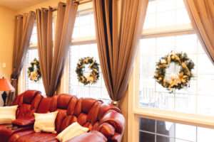A green wreath with a gold ribbon running around, and gold, clear, and white ornaments hanging on a window next to taupe drapes, above a reddish-brown sofa.