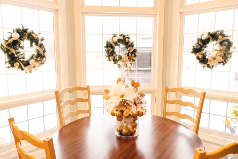 A green wreath with a gold ribbon running around, and gold, clear, and white ornaments hanging on a window behind a wooden table with a white and gold floral arrangement in the center.