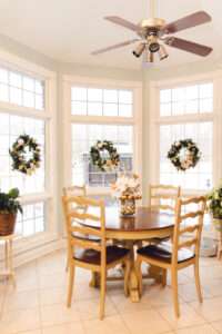 3 green wreaths with gold ribbon running around, and gold, clear, and white ornaments are centered on 3 windows behind a wooden table with a white and gold floral arrangement in the center.
