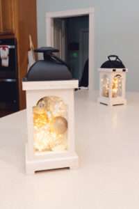 White lanterns with matte black handles, filled with white, gold, and clear ornaments with lights inside.