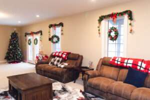 Greenery with red and green ornaments, and a green wreath with red and green ornaments, hanging over 2 windows behind some dark from furniture.