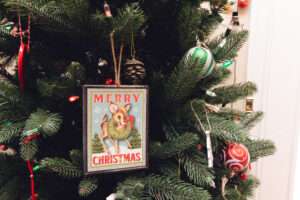 A close up of a Merry Christmas ornament on a green Christmas tree with red and green ornaments surrounding it.